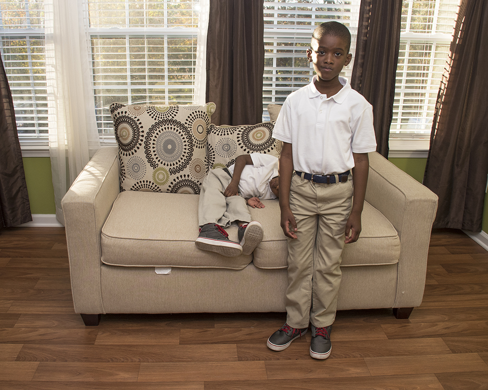 Two young brothers posing for a picture, one standnig up the other laying on a couch